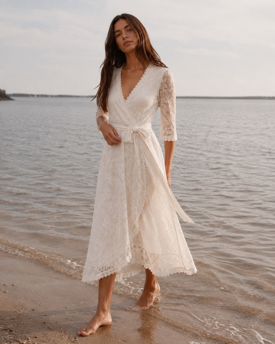 Petite model in an ivory lace wrap midi dress walking barefoot along the shoreline.
