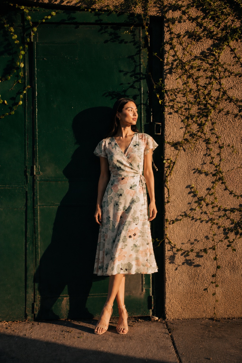 Petite model in a floral wrap midi dress against a green door at golden hour.