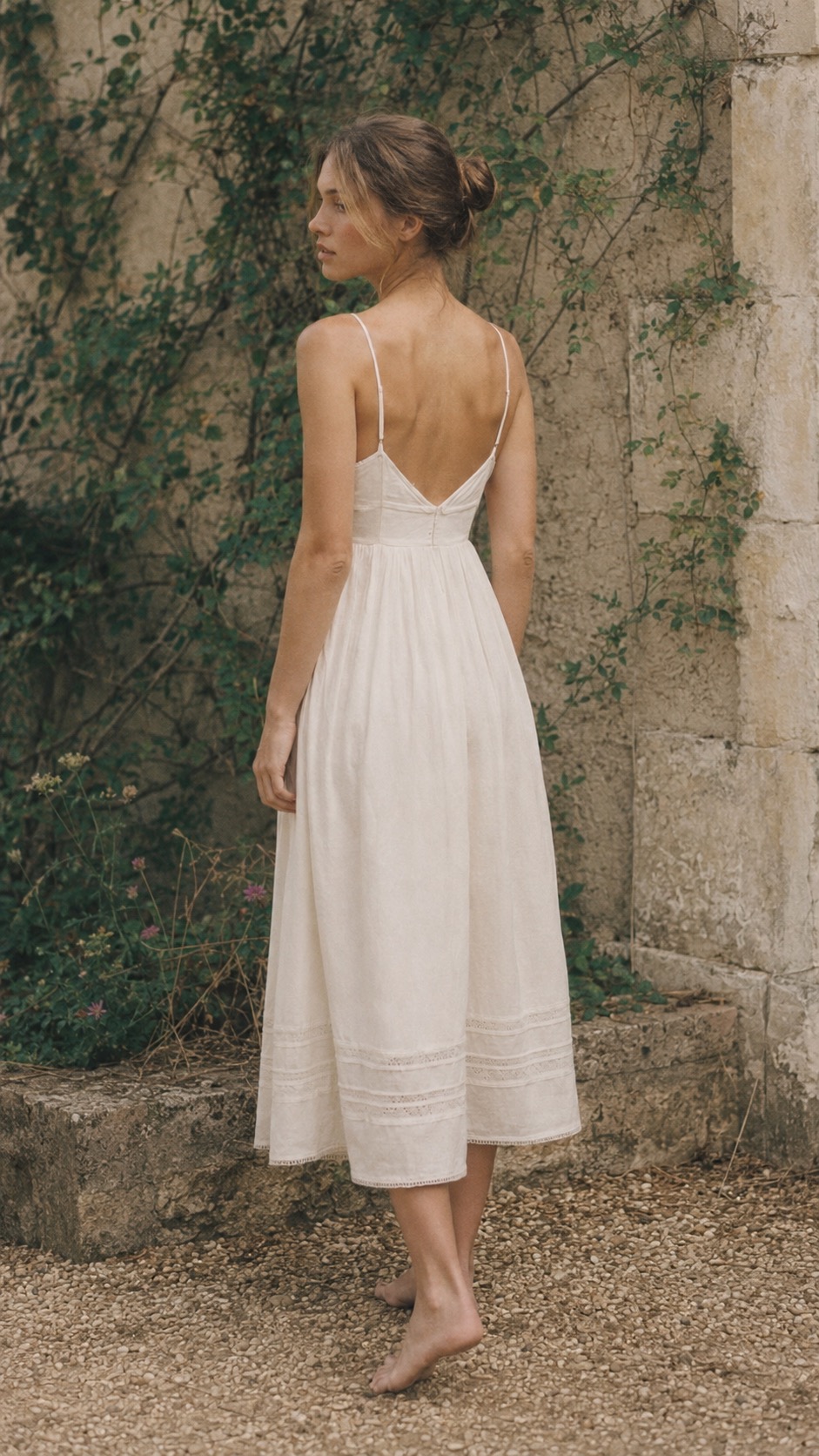 Petite woman in an ivory linen slip dress with lace hem, seen from behind against an ivy-covered stone wall.
