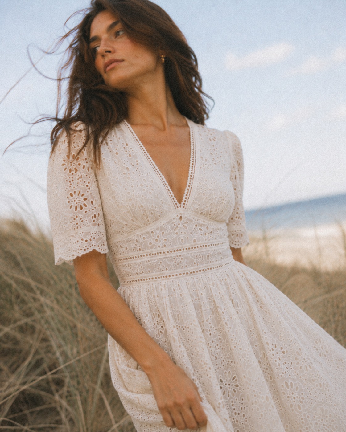Petite model in an ivory eyelet midi dress on a windswept beach at golden hour.