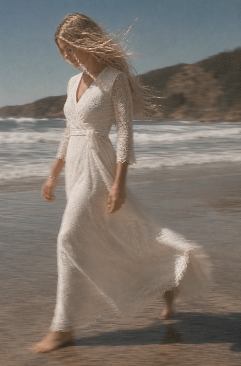Woman walking barefoot along the shoreline in a floor-length ivory lace maxi dress with three-quarter sleeves.