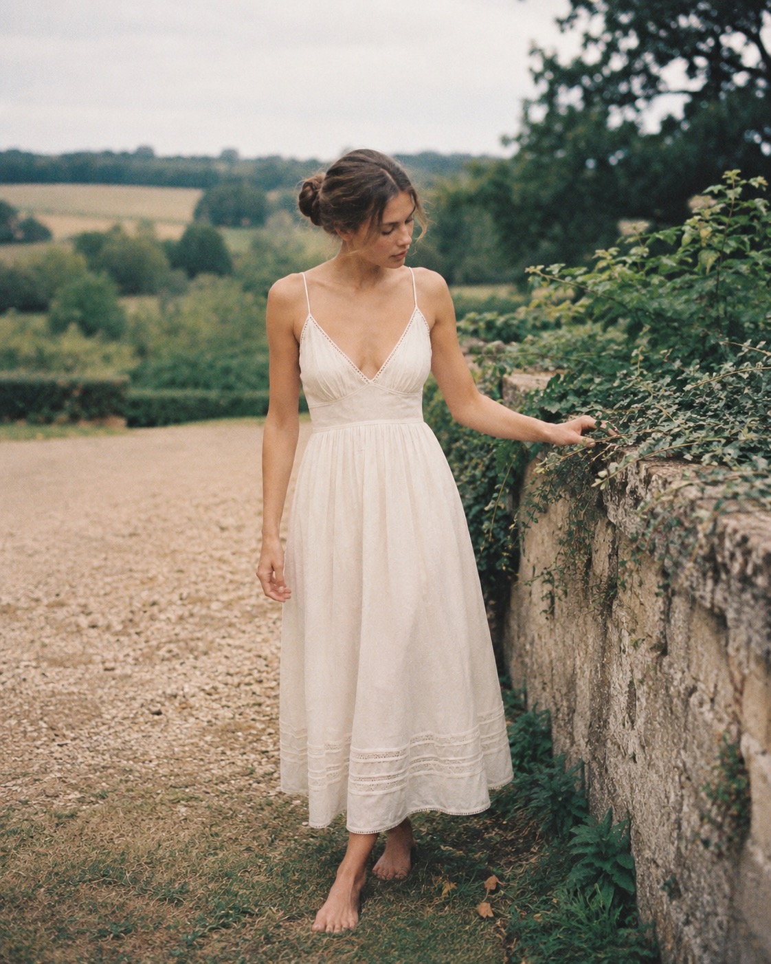 Woman in an ivory linen slip dress with lace hem standing by a countryside garden wall