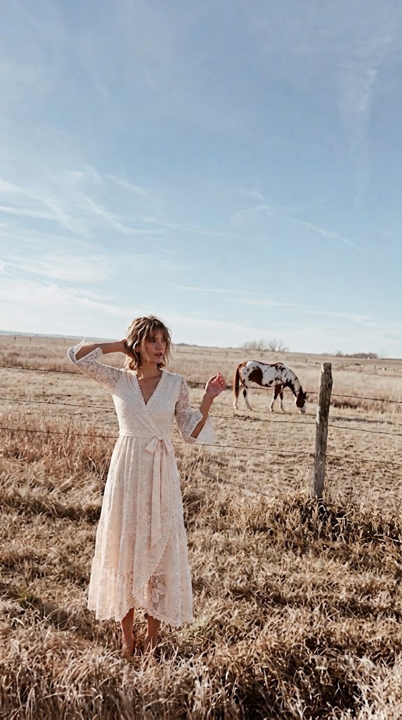 Woman in an ivory lace wrap dress in a grassy pasture beside a fence and grazing horse