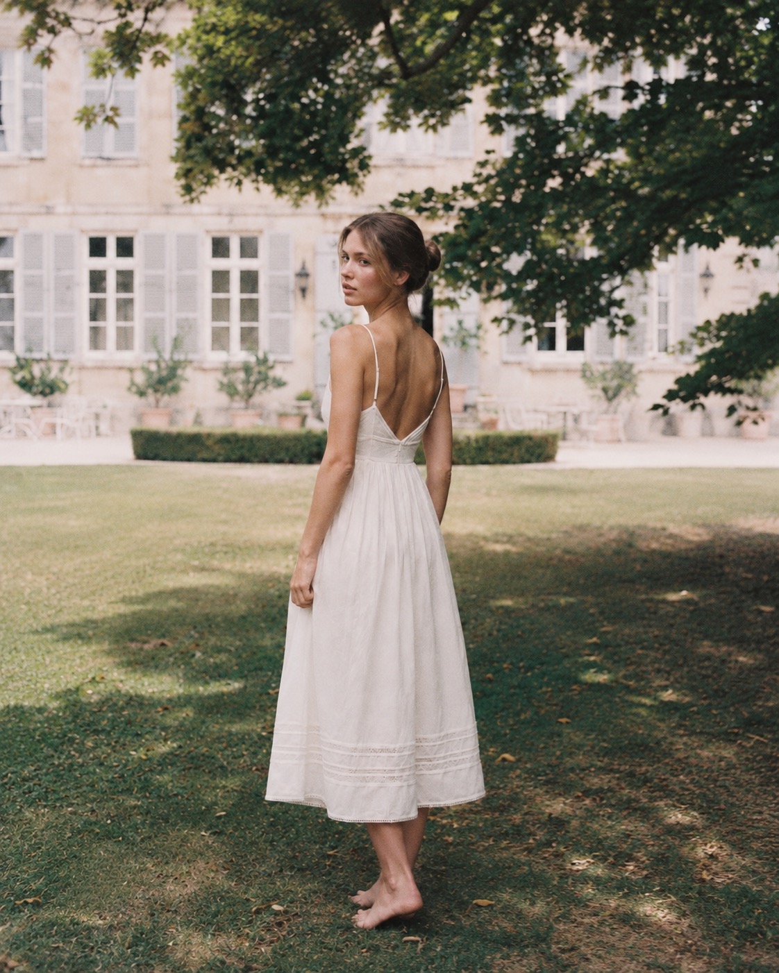 Woman in an ivory linen slip dress with lace hem, seen from behind on a château lawn