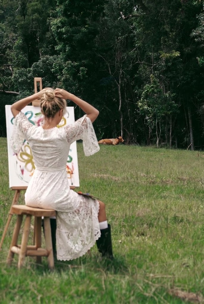 Woman in an ivory lace dress and black boots painting at an easel in a green pasture with a cow resting by the trees